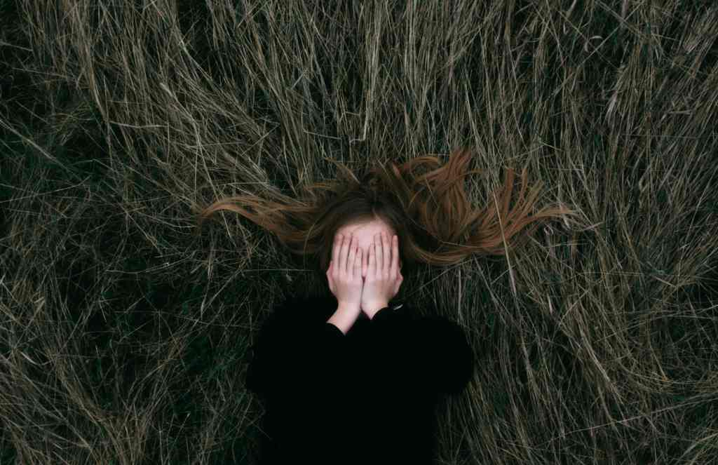 Overhead shot of a girl lying in tall dry grass covering her face with hands, outdoors nature scene.